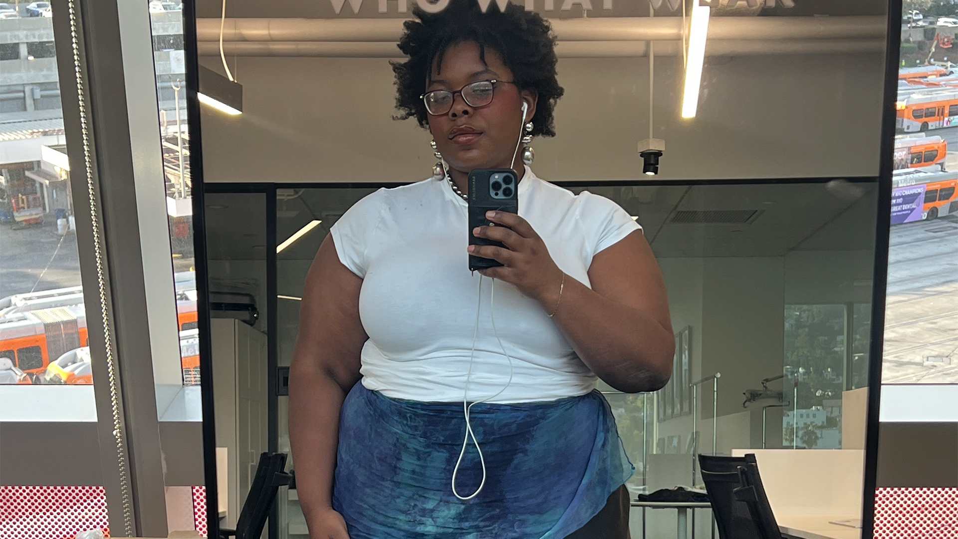 Chichi takes mirror selfie in white tshirt, black slip skirt, blue scarf around the waist, and silver shoes and jewelry