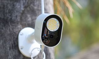 A white wireless outdoor security camera with built-in LED floodlight and Wi-Fi antenna, wall-mounted on a wooden post.