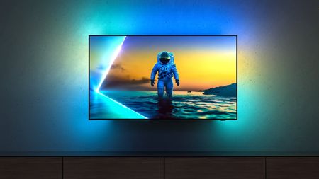 A wall-mounted 2025 Philips OLED TV with Ambilight. There is an astronaut on the screen and blue, green and yellow ambient light is being thrown onto the wall behind the display.