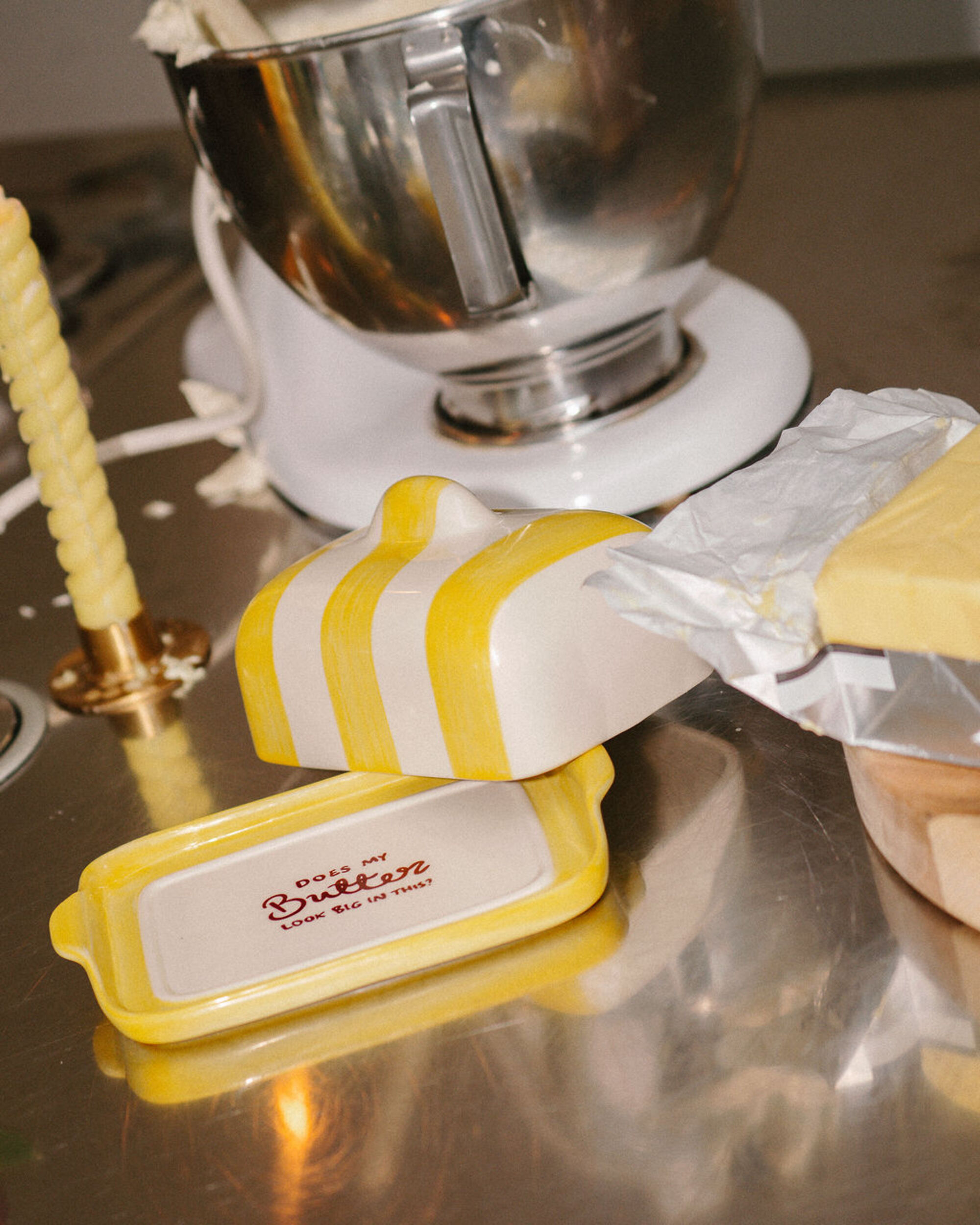 Image of a yellow and white striped butter dish on a metal kitchen countertop with a stainless steel mixing bowl in the background.
