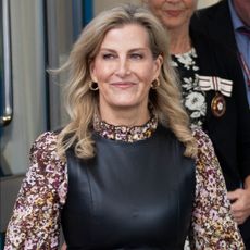 Duchess Sophie leaving a building wearing a black leather dress over a patterned floral blouse and smiling