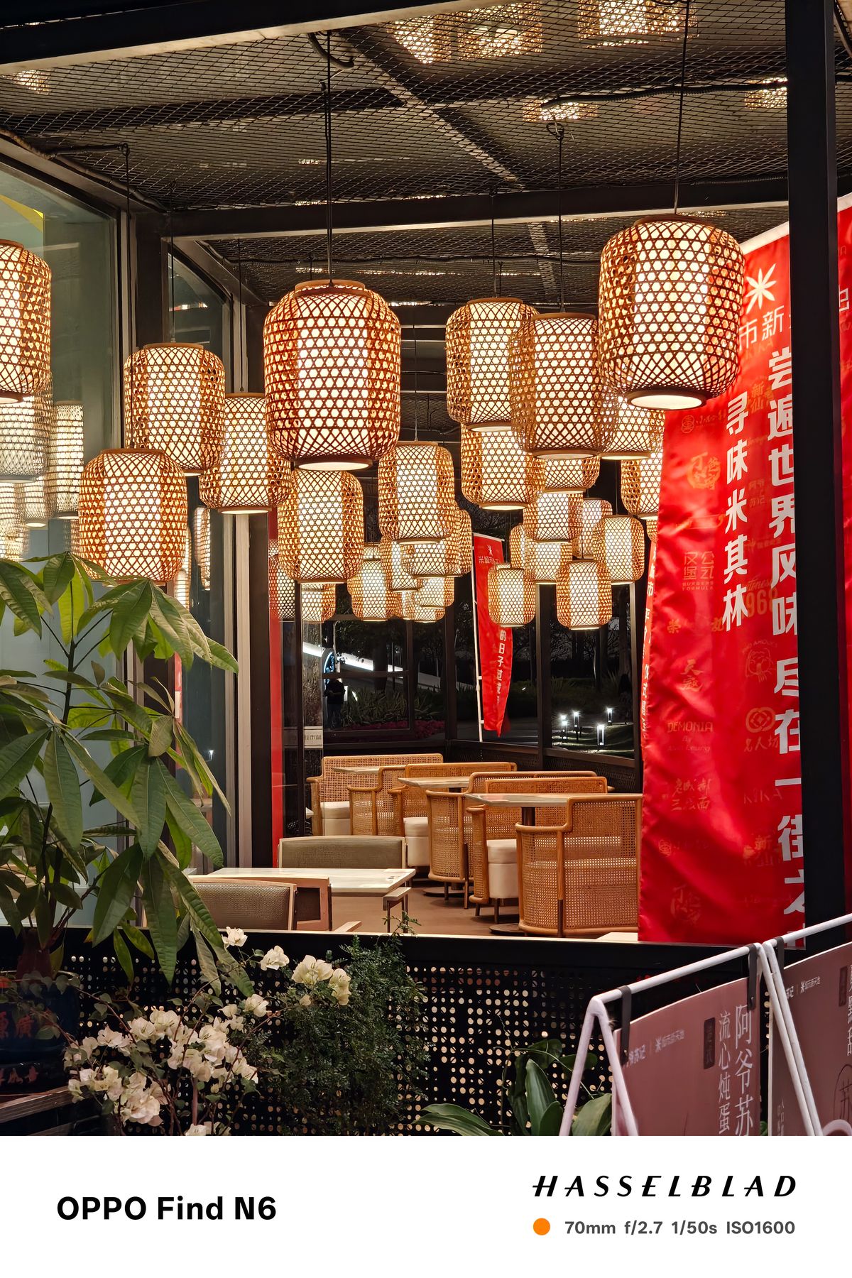 A wide interior shot of a restaurant or lounge featuring dozens of large, woven bamboo lanterns hanging from the ceiling at different heights. Below the glowing lanterns are several woven armchairs and tables, with a large red banner featuring white Chinese text on the right.
