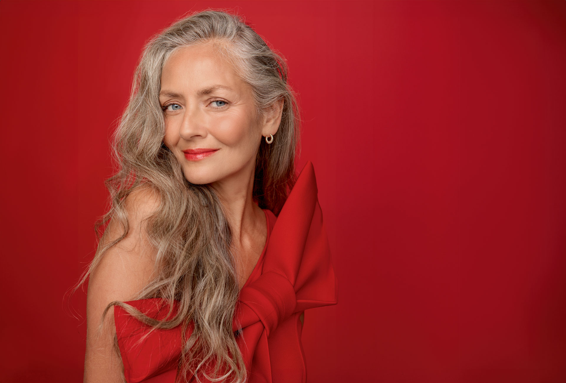 A woman with long grey and blonde hair, styled in side parted long flowing waves. She is wearing a red dress, red lipstick and gold earrings