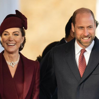 Kate Middleton wearing a maroon coat and hat and smiling next to Prince William wearing a dark coat and a red tie 