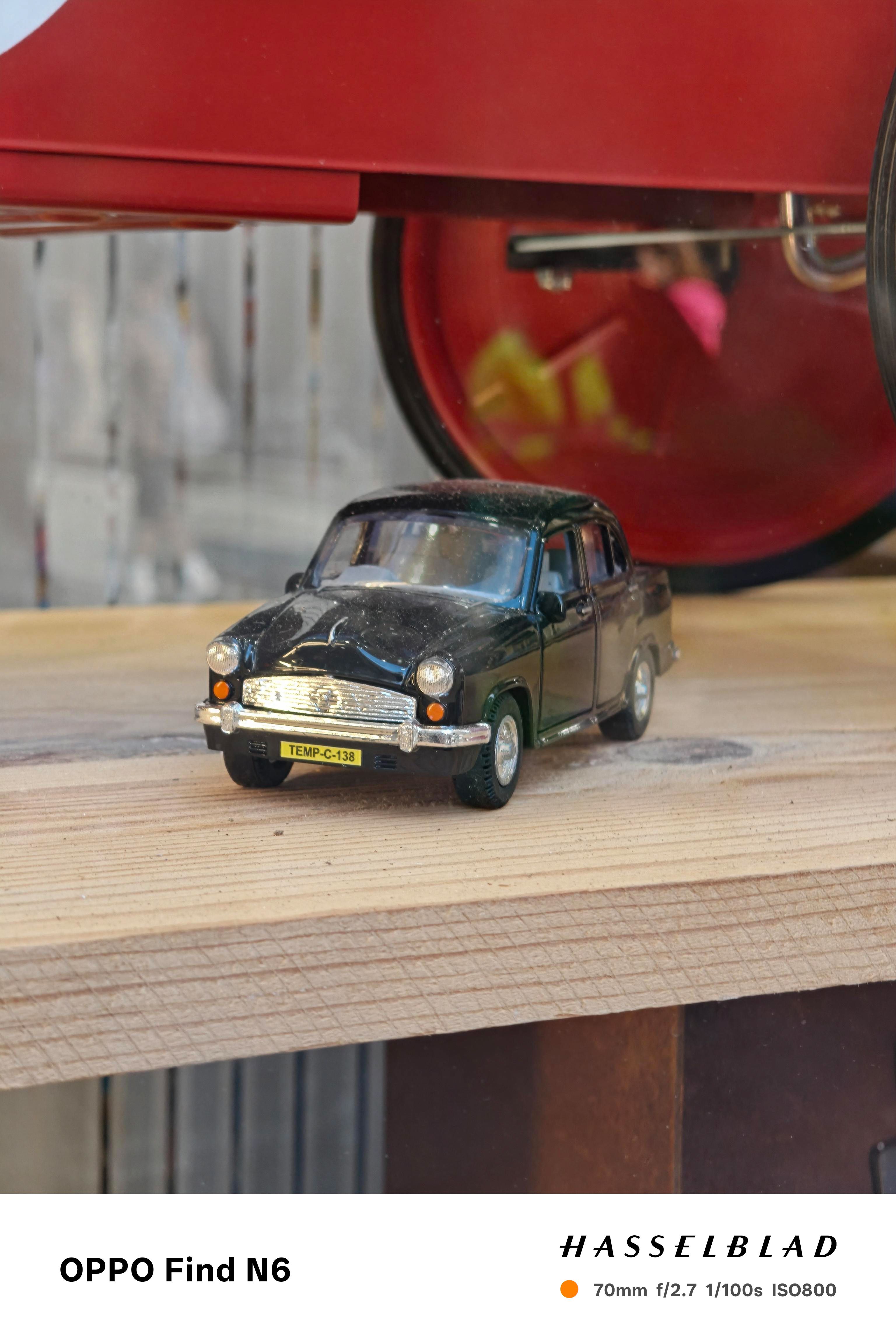 A close-up photograph of a vintage-style black toy car with silver chrome bumpers and a yellow license plate that reads "TEMP-C-138." The car is resting on a light wooden shelf, with a soft, blurred reflection visible in the glass behind it.