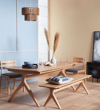 Light wood wishbone style dining table and bench, tiered wood chandelier, and light blue and sand walls, and pampas grass in vases on table.