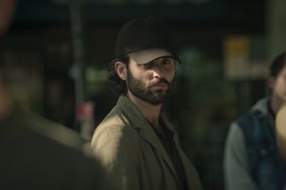 Penn Badgley as Joe Goldberg, wearing a baseball cap while standing in a crowd, in episode 406 of You.