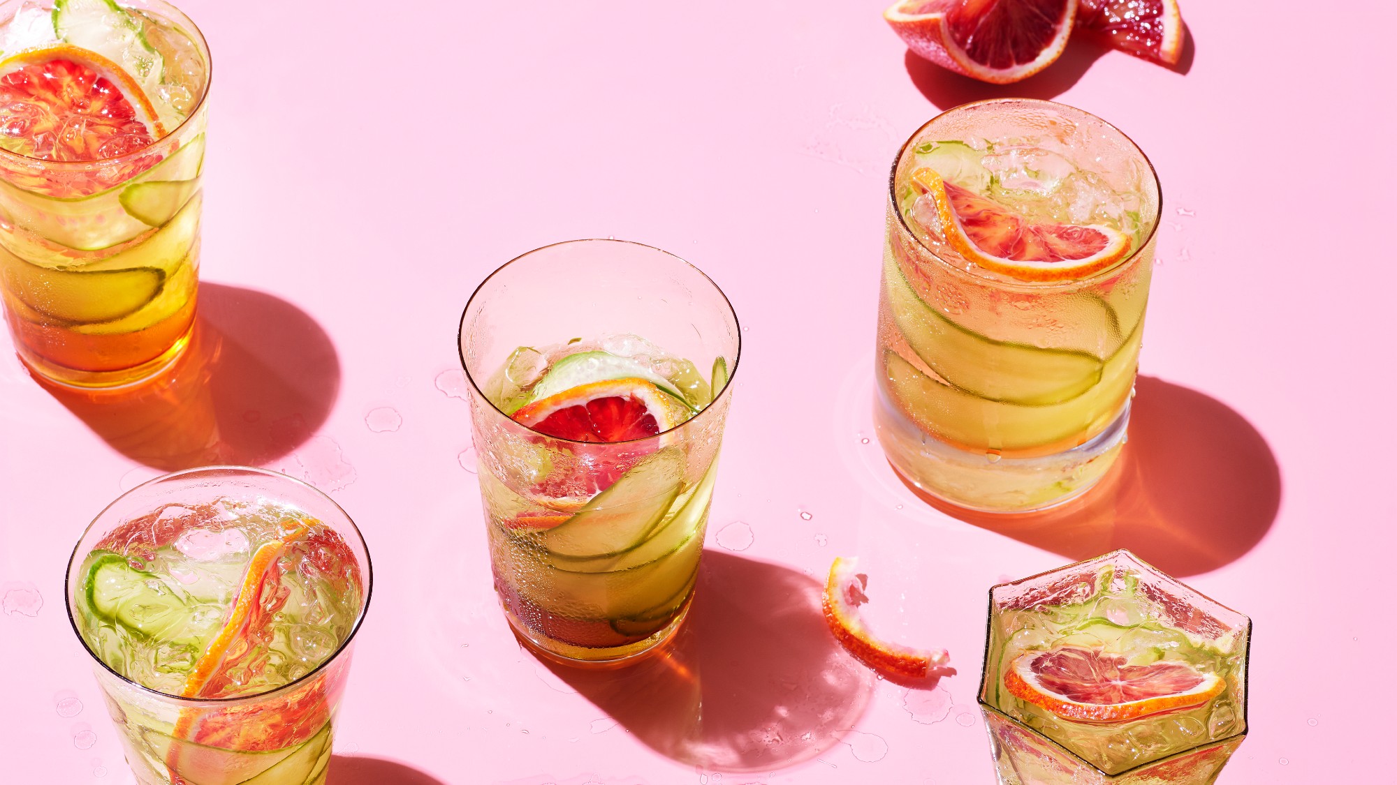 3/4 overhead shot of five glasses with a cucumber-grapefruit drink inside them. the backdrop is bright pink. 