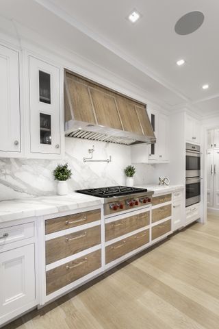 A bright white kitchen with white cabinets, wooden drawers, wood flooring, and marble countertops