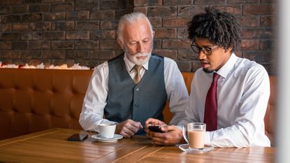 An older man and a younger man look at a phone together in a coffee shop.