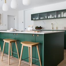 green and white kitchen with green island, white worktop, narrow glass fronted wall units, oak wood bar stools