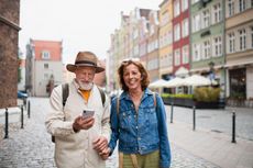 2JBFPXD Portrait of happy senior couple tourists smiling, holding hands, using smartphone outdoors in historic town