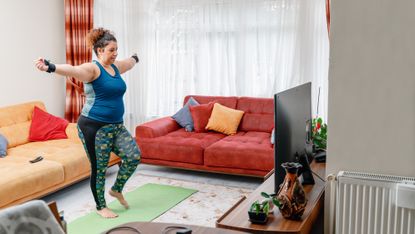 A woman exercises at home on a yoga mat wearing wrist weights. She is marching on the spot, with her arms held out to the side, looking at the television in front of her. Behind her are two couches with scattered cushions and a long white curtain covering a window. 