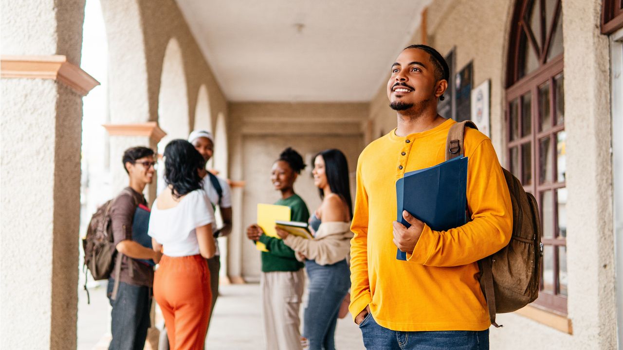 A college student looks into the distance while standing outside a classroom and holding books.