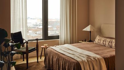Image of a beige bedroom with minimalist brown bedding and a beige throw at the end of the bed. There is a black and wood accent chair in the corner.
