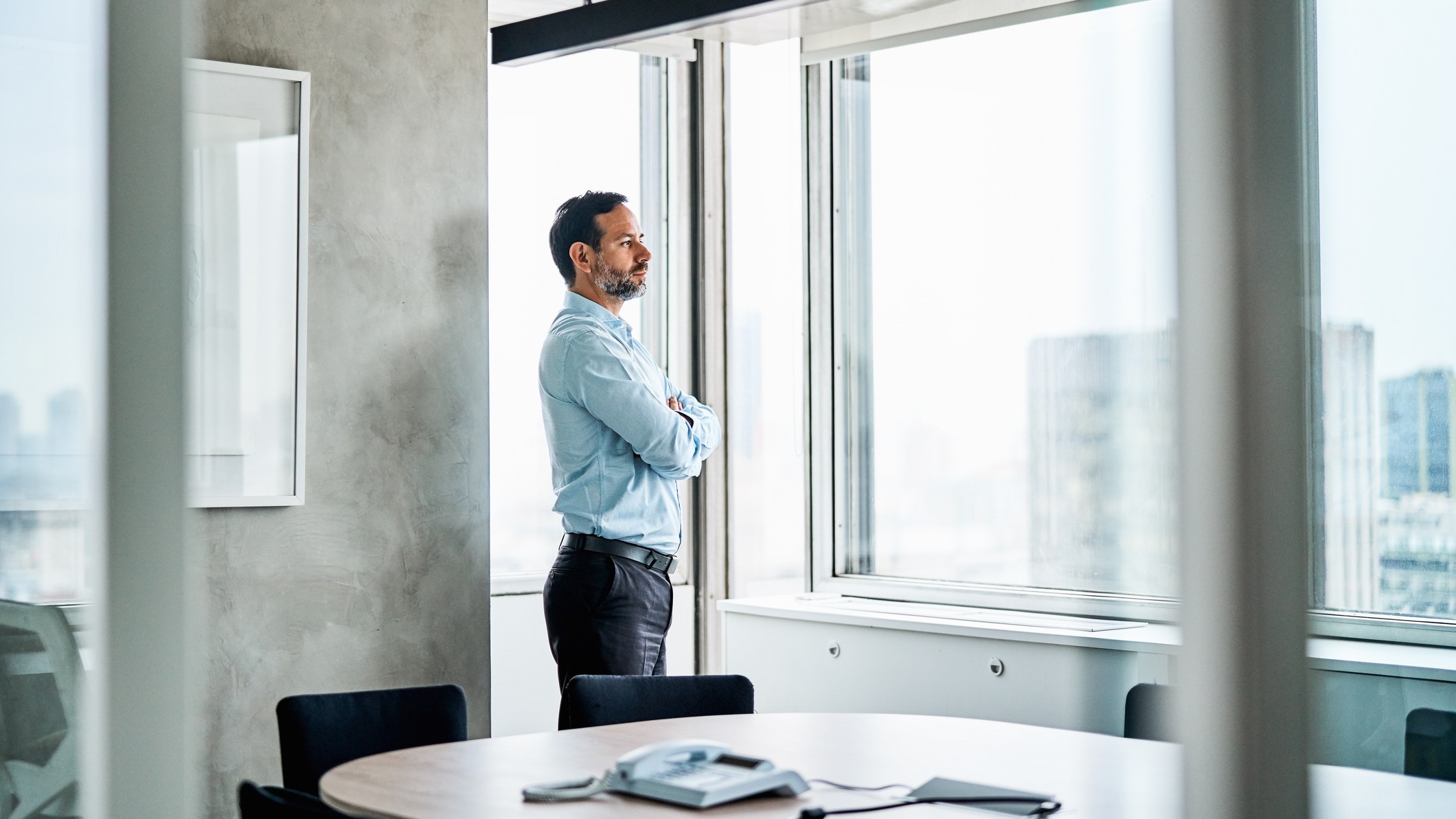 A man in business clothes looks out a window in an empty conference room. 