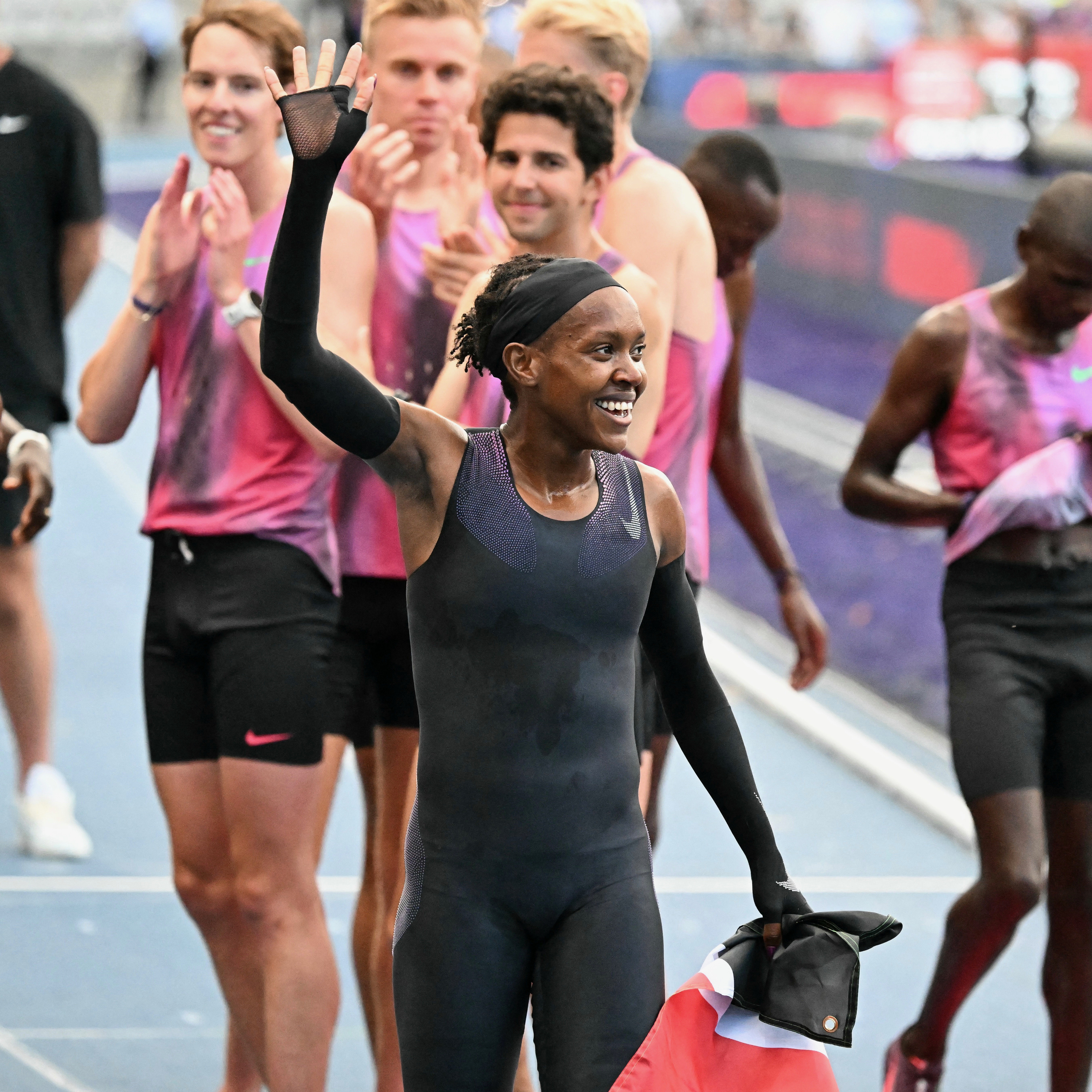 Faith Kipyegon raises her arm in triumph after her race, surrounded by pacemakers in matching pink and black Nike gear at Paris’ Stade Charléty.