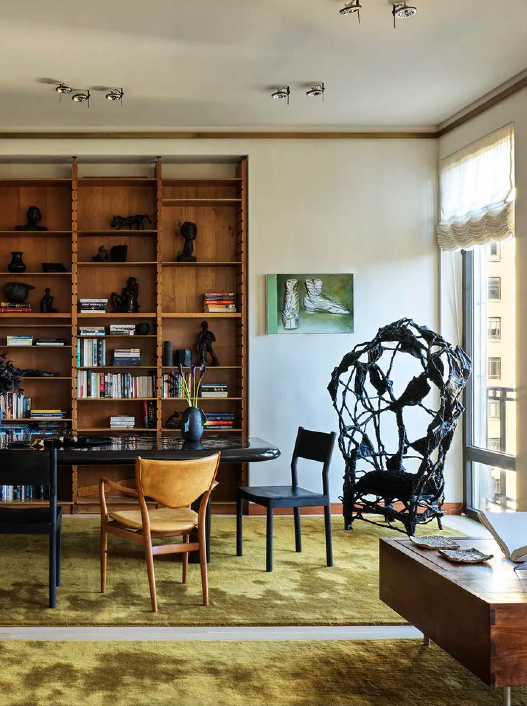 A large wooden shelving unit filled with books and decorative objects in front of a black oval dining table with black and wood dining chairs with a black sculpture in the corner of the room