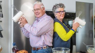 Prue and her husband John in their kitchen both holding electric whisks up in front of them