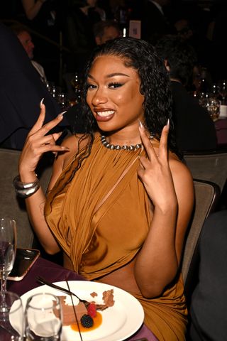 Megan Thee Stallion sits at a table at a dinner in a brown dress and long nails.