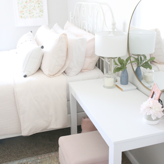 Light bright, feminine bedroom with desk-cum-dressing table, table lamp, and large round wall mirror.
