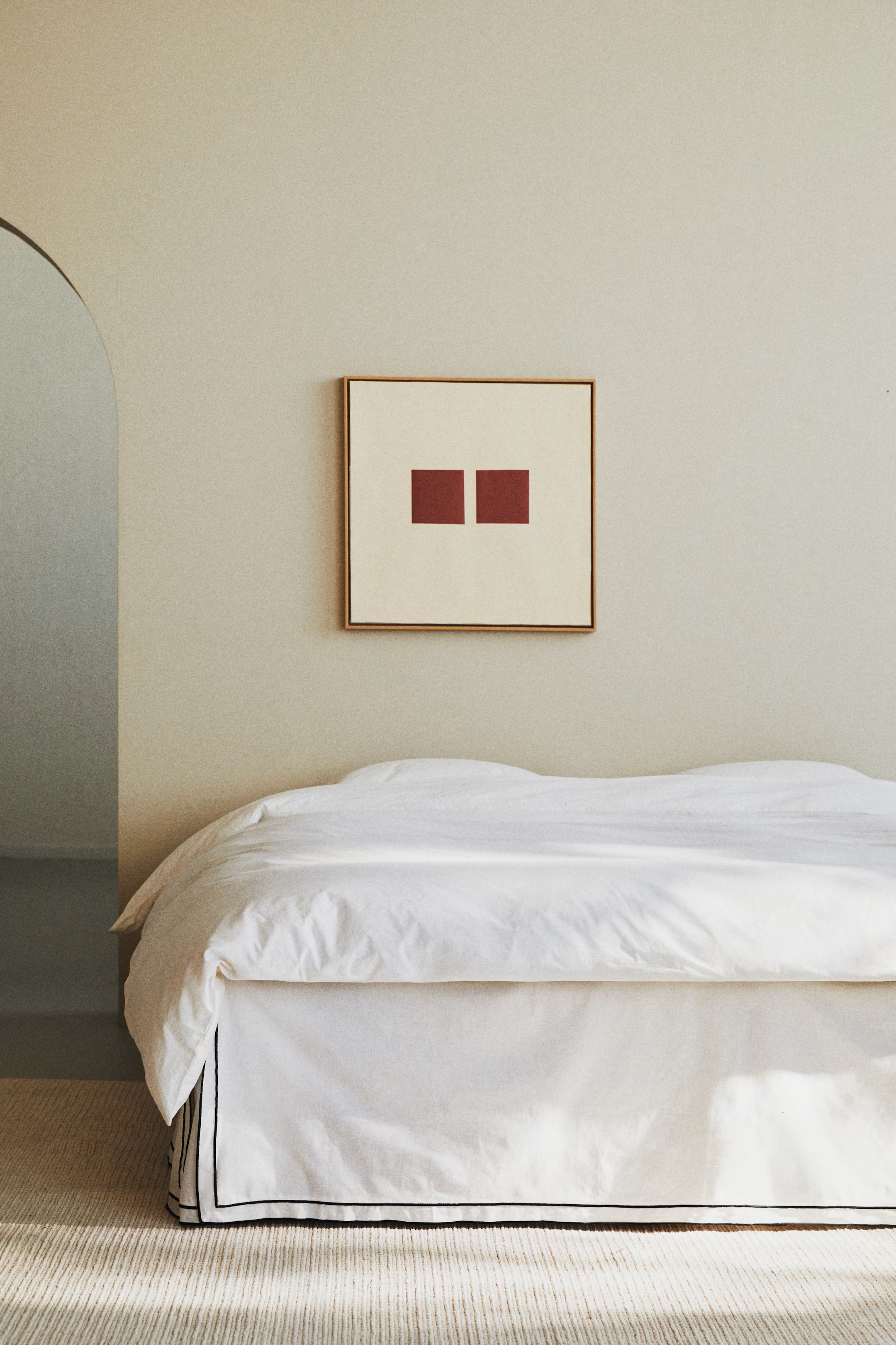 Image of a minimalist white bed with a white bed valance that has navy trim. The bedroom is also minimal, with beige walls and a photo hanging of two small red squares.