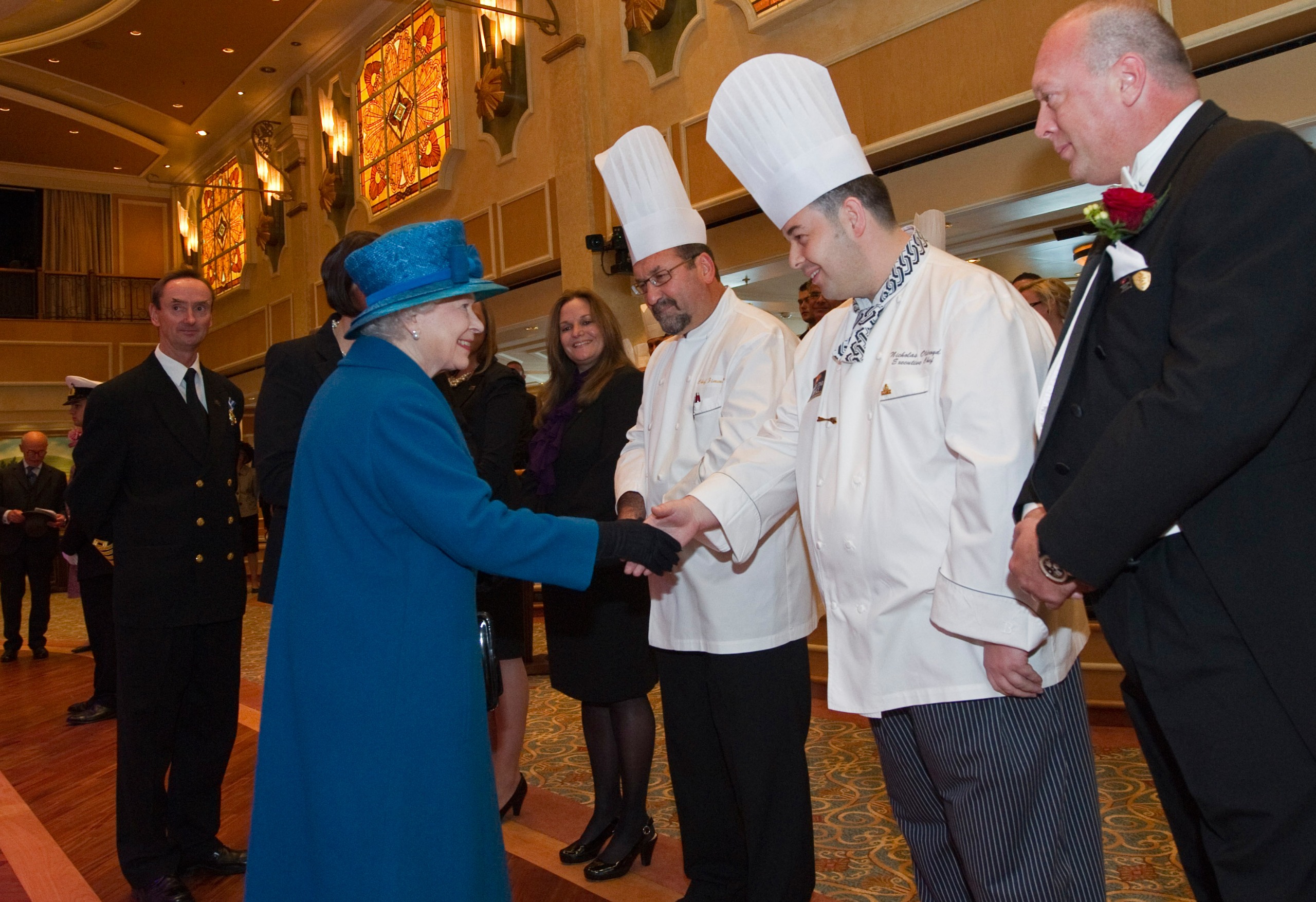 Queen Elizabeth wearing a blue coat and hat shaking hands with a chef on a cruise ship