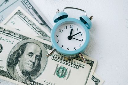 Blue vintage alarm clock lying over four one-hundred-dollar bills on white textured surface.