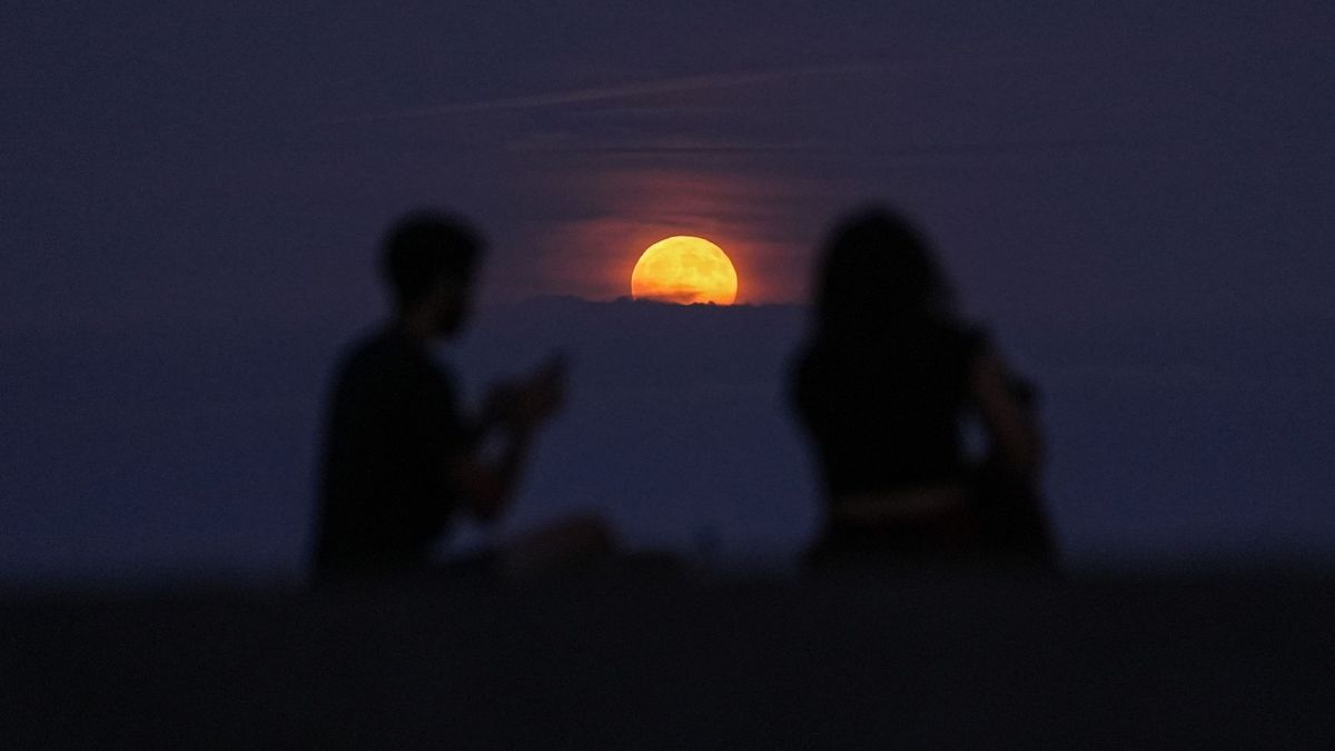 Una luna llena de color naranja se eleva sobre un banco de nubes en un oscuro cielo nocturno entre las siluetas de dos espectadores.