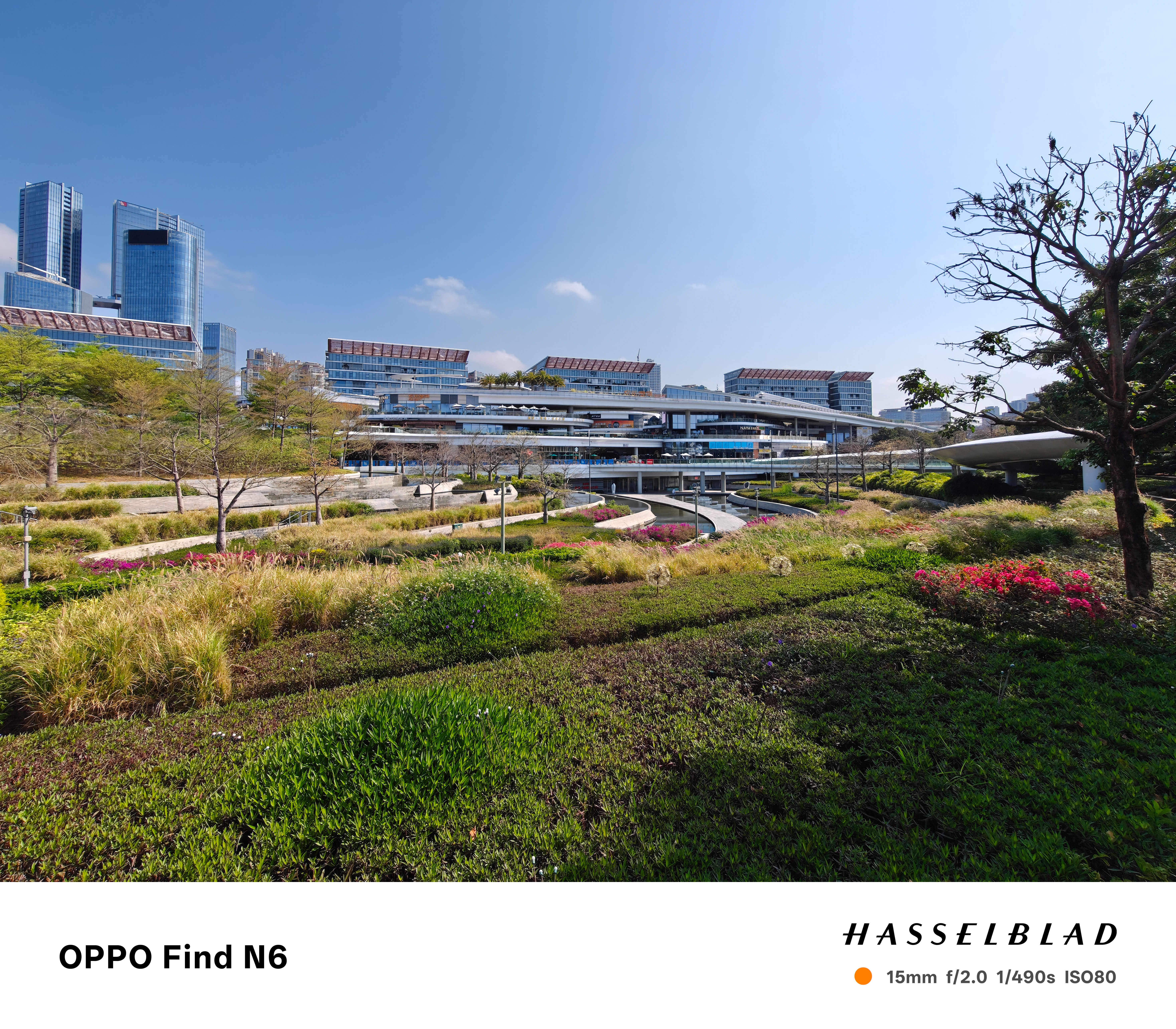 An ultra-wide-angle landscape photograph of a modern, multi-tiered outdoor shopping complex with white facades and glass railings. In the foreground, a lush green park features terraced lawns, ornamental grasses, and pink flower beds under a clear blue sky.