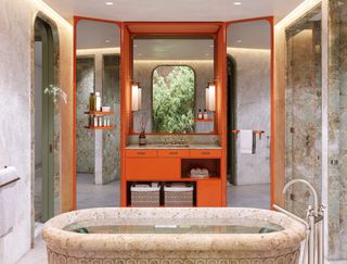 The bathroom of a luxury hotel room features marble and stone finishes contrasting the pastel-shaded red and mirror surfaces of its cabinet.