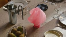 A close-up image of a table set with a beige table cloth, wine glasses, a bowl of pears, and a chrome water jug. There is a pink, swirly table lamp in the center of the table.