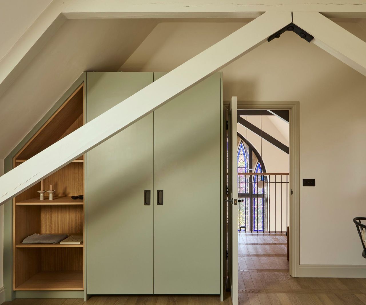 A vaulted bedroom with white timber roof beams, sage green fitted wardrobe, open oak shelving and a doorway revealing a stained glass window beyond.