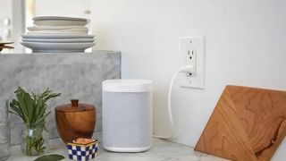 A white Sonos One speaker plugged into a wall socket on a marble surface next to a chopping board.