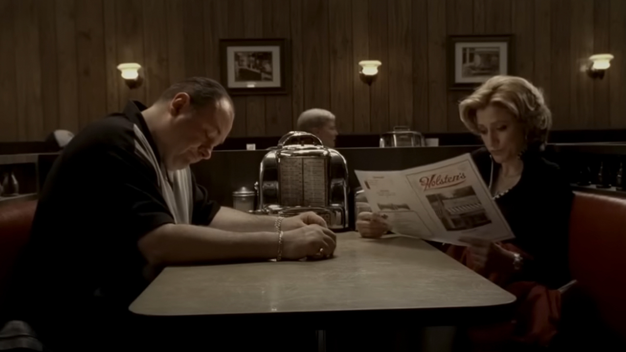 James Gandolfini on the left, sitting in a booth with Edie Falco as they both look at menus in The Sopranos.