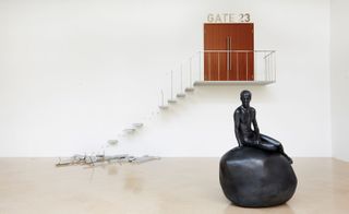 A white wall with a broken staircase going up it to a brown door and a black sculpture of a man sitting on a rock below it.