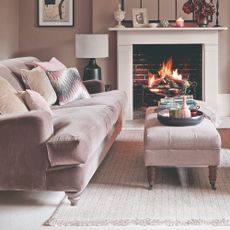 A living room with a pink sofa and a matching ottoman for a coffee table with the fireplace lit in the background