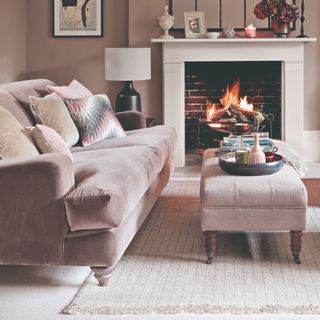 A living room with a pink sofa and a matching ottoman for a coffee table with the fireplace lit in the background