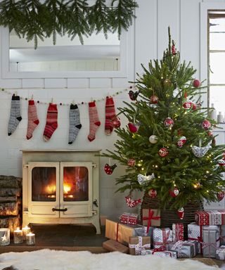 Country Christmas living room with red and blue knitted stockings hung up over a woodburning stove, next to a christmas tree with wrapped presents underneath