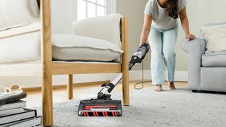 Woman using a shark cordless stick vacuum in her home vacuuming under a chair.