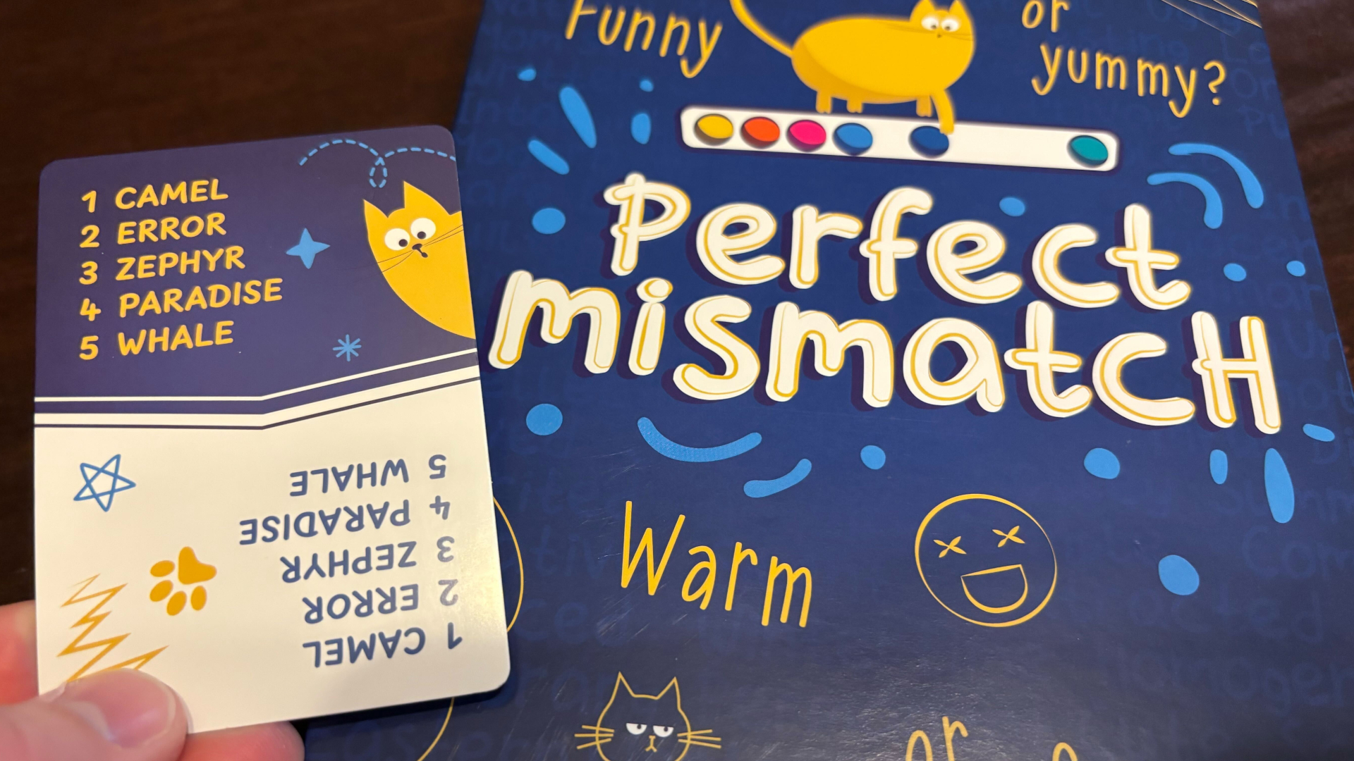 A hand holds a card with numerous words against the Perfect Mismatch box, which sits on a dark wooden surface