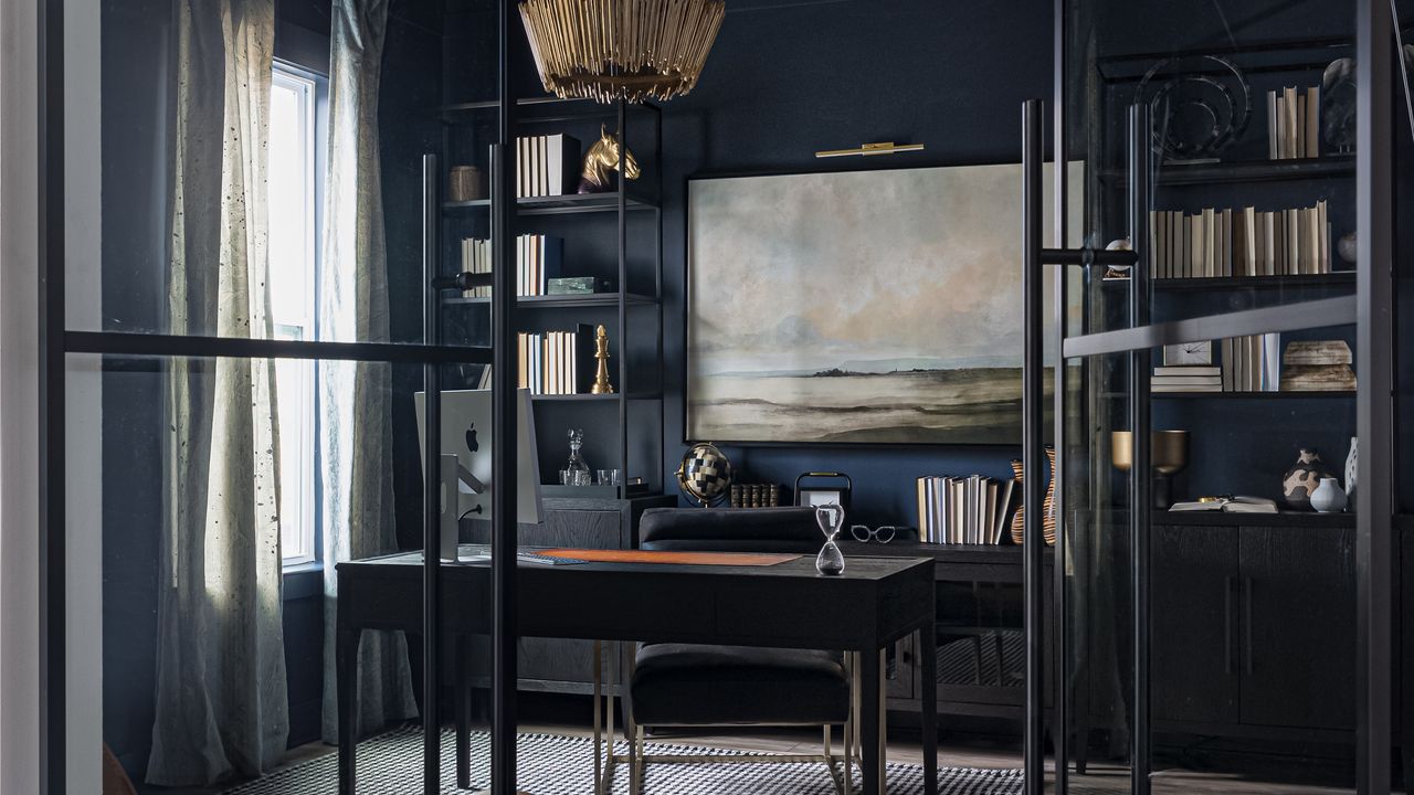 Image of a traditional style home office that is painted in a navy blue paint color. There is a slim black desk and black desk chair and two slim black bookshelves on either side of the back wall. There is a light, landscape painting in between the shelving units.