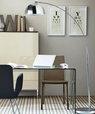 A modern home office with light gray walls with two framed cream wall art prints, a tall silver lamp, a cream white unit with brown books, and a desk with a laptop and two chairs either side