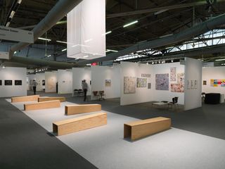 An exhibition space with eight booths standing with artwork inside. A row of wood benches in the foreground.