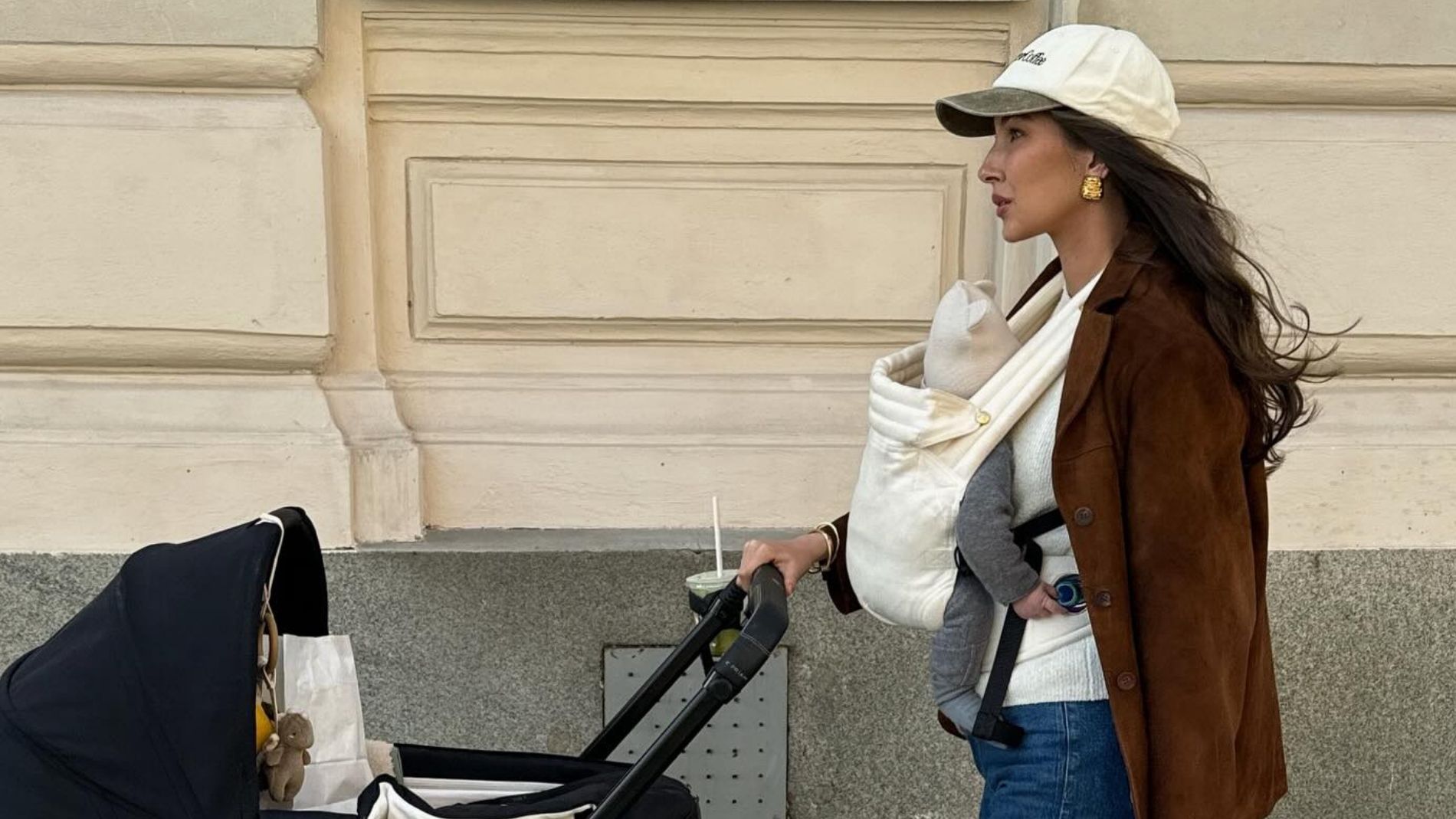 A new mom carries her baby while wearing a brown suede jacket with jeans and Adidas sneakers.