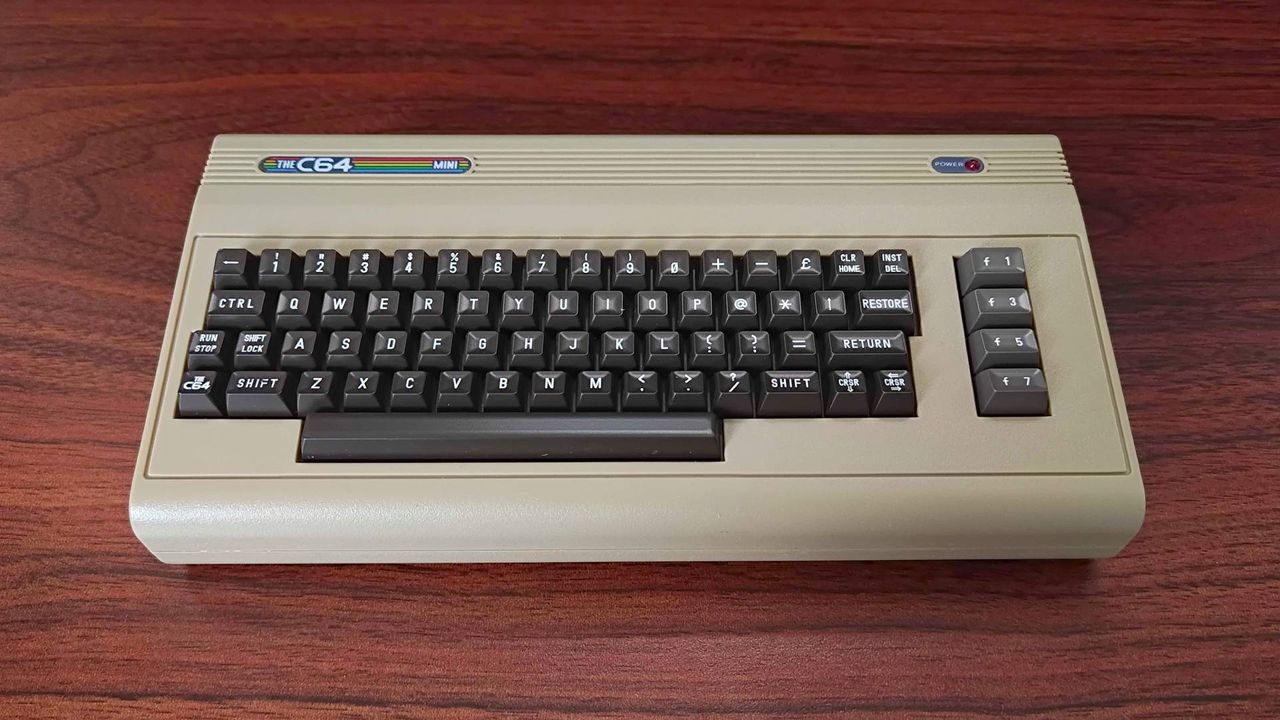 Front of The C64 Mini on woodgrain desk with faux keyboard, power light, and badge in view.