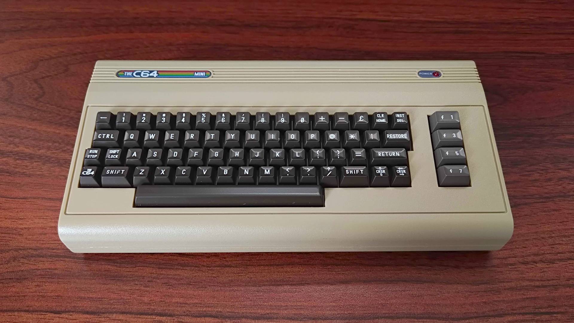 Front of The C64 Mini on woodgrain desk with faux keyboard, power light, and badge in view.