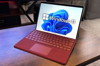 Windows 11 logo superimposed onto the screen of a Microsoft Surface Pro 9 with a red keyboard seated on a wooden desk