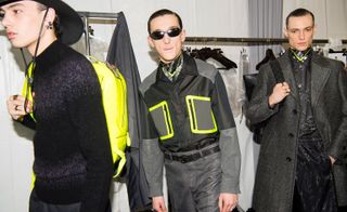 Three male models wearing looks from Louis Vuitton's collection. One model is wearing a black cowboy hat, black jumper with two lighter coloured stipes and is holding a yellow backpack. Next to him is a model wearing sunglasses, a black, grey and yellow shirt, necklace, grey trousers and belt. And the third model is wearing a dark coloured piece, necklace, grey coat and is holding a bag with the signature Louis Vuitton pattern