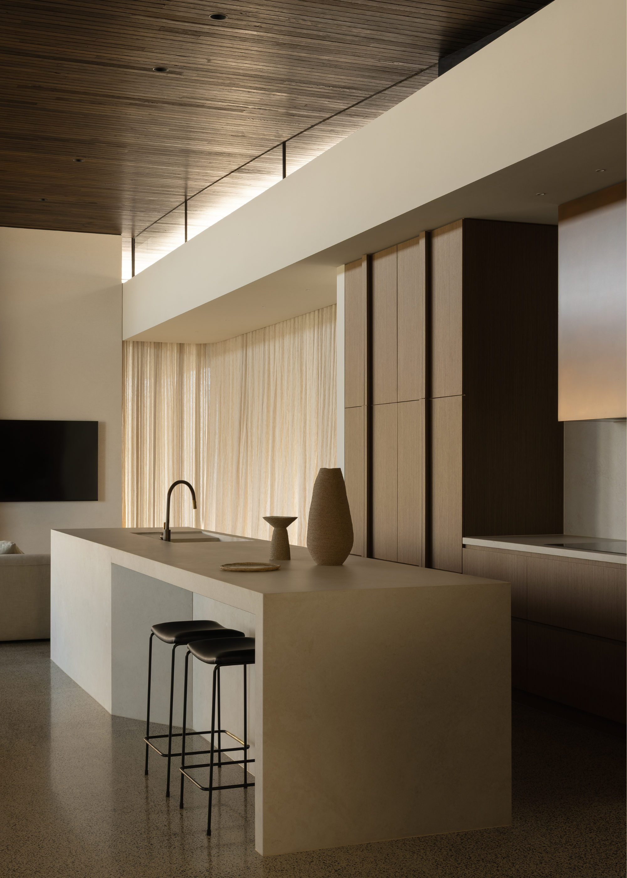 A minimalist kitchen with a stone island, beige ceramics, black stools, and wood cabinets